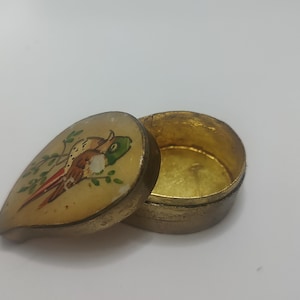 以下が含まれることがあります： 葉の形をした蓋が付いた、小さな丸い金色の宝箱。蓋には、茶色、赤、緑の色合いの鳥が描かれています。