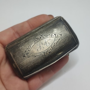 May include: Antique silver rectangular box with ornate engraved floral patterns. The box features a central oval cartouche with the engraved initials "J A". The box is held in a hand.