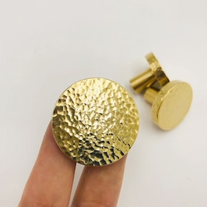 Op de afbeelding: Twee gouden ronde kastknoppen met een gehamerd oppervlak. De knoppen hebben een getextureerd oppervlak en worden getoond op een witte achtergrond.