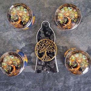 May include: Five pieces of jewellery are displayed on a grey surface. Four round plugs feature a colourful tree design. A gold-coloured tree of life pendant is attached to a black and silver pendant.