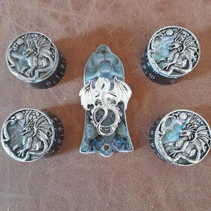 Könnte beinhalten: Fünf silberne Drachenknöpfe mit schwarzen und weißen Abalone-Schalen-Einlagen. Die Knöpfe sind rund und haben ein erhabenes Design eines Drachen mit Flügeln. Die Knöpfe sind in einem Kreis um einen größeren, rechteckigen Knopf mit einem ähnlichen Drachen-Design angeordnet.