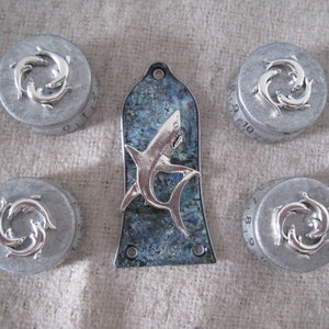 Pode incluir: Quatro caixas redondas prateadas com um desenho de três golfinhos nadando em círculo. Uma quinta caixa é um protetor de guitarra com um design azul e branco e um tubarão prateado no centro.