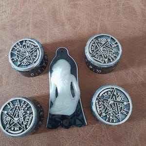 Könnte beinhalten: Vier schwarze und silberne Knöpfe mit einem fünfzackigen Sternmuster und den Worten "GRAM", "NOL", "TRA" und "MATON" um den Stern herum. Die Knöpfe befinden sich auf einem schwarzen und weißen Gitarrenplektrum mit einer weißen Figur in der Mitte.