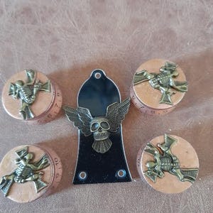 Puede incluir: Cinco pomos decorativos con un diseño de calavera y huesos cruzados. Los pomos son de metal y tienen un acabado marrón. Los pomos están dispuestos en círculo alrededor de una placa de protección de guitarra negra.
