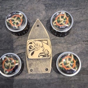 Könnte beinhalten: Eine Sammlung von Gitarrenteilen, darunter vier schwarze Knöpfe mit silbernen Rändern und eine dekorative Platte. Die Knöpfe weisen ein farbenfrohes Design auf, und die Platte hat ein cremefarbenes Jugendstil-Bild.