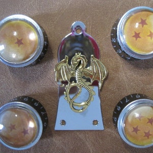 Puede incluir: Un conjunto de cinco perillas de guitarra con acabado plateado y una parte superior transparente en forma de cúpula. Las perillas presentan un diseño que recuerda a una bola de dragón, con estrellas en el interior. También se incluye una placa de guitarra dorada con forma de dragón.