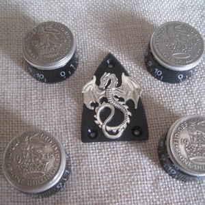 Puede incluir: Cinco pomos negros con monedas de plata en la parte superior. Las monedas son monedas británicas de una chelín de 1919 y 1948. Los pomos tienen números del 0 al 10 impresos en ellos. En el centro hay una placa de guitarra negra con un diseño de dragón plateado.