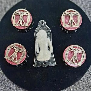 Könnte beinhalten: Eine Sammlung dekorativer Knöpfe und ein Anhänger. Die Knöpfe zeigen ein goldfarbenes Vitruvianischer-Mann-Design auf rosa Hintergrund, mit schwarzen Zahlen am Rand. Ein weißer, stilisierter Figurenanhänger ist ebenfalls vorhanden.