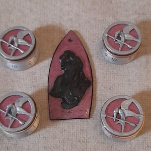 May include: Five round, silver-coloured, metal knobs with a pink background and a black and silver bird design. The knobs have numbers 10, 9, 10, 10, and 10 printed on them. A pink leather patch with a black silhouette of a person is in the centre of the image.