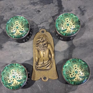 Puede incluir: Cuatro perillas verdes y doradas con un diseño de árbol celta y un colgante de color latón con la figura de una mujer. Las perillas tienen bases negras con marcas numéricas blancas. El colgante tiene un agujero en la parte superior.