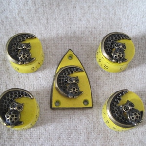 May include: Five yellow knobs with black numbers and an owl design. The knobs have a crescent moon shape with an owl perched on the moon. The numbers on the knobs range from 0 to 10.