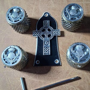 May include: A set of silver-toned guitar knobs and a black headstock plate with a Celtic cross design. The knobs feature a thistle emblem. An Allen wrench is also included.