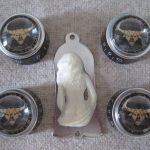 Op de afbeelding: Vier zilveren knoppen met zwarte stierenkopontwerpen en de nummers 6-10 erop gedrukt. Een witte plastic gitaarplectrum in de vorm van een vrouw bevindt zich in het midden.