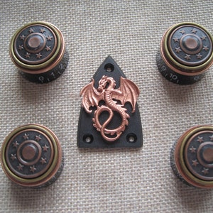 Puede incluir: Conjunto de cinco botones de guitarra de cobre y negro con un diseño de dragón de cobre en una placa de protección negra. Los botones tienen un diseño circular con un anillo de estrellas alrededor del borde.