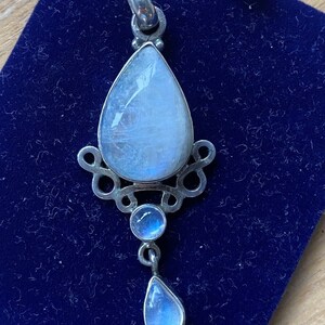 May include: A silver pendant featuring a large teardrop-shaped moonstone, with two smaller moonstones dangling beneath. The pendant is displayed on a blue velvet background.