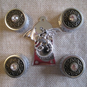 Könnte beinhalten: Fünf silberne und goldene Knöpfe mit einem Sternmuster und einem klaren Strassstein in der Mitte. Die Knöpfe sind von einem silbernen Drachenanhänger umgeben.