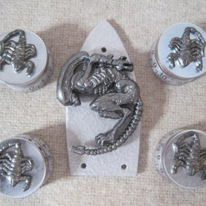 Puede incluir: Cinco pomos de metal de color plateado con un diseño de escorpión. Los pomos están numerados del 1 al 10. Una criatura extraterrestre de metal plateado se encuentra en el centro de la imagen.