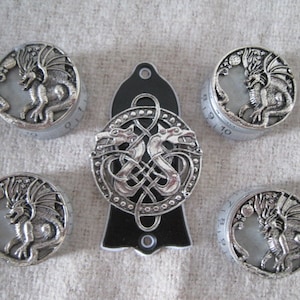 Könnte beinhalten: Eine Sammlung von silberfarbenen Metallknöpfen und einer zentralen Platte, alle mit Drachendesigns. Die Knöpfe sind kreisförmig mit einem Drachenrelief, während die Platte ein keltisches Knotenmuster mit zwei Drachen aufweist. Der Hintergrund ist ein neutraler Stoff.