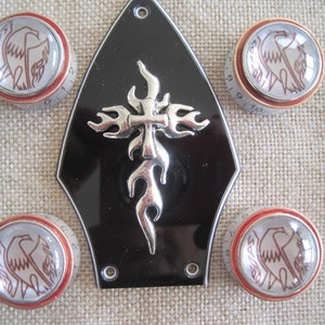 Puede incluir: Un pickguard de guitarra negro con un diseño de cruz plateada con llamas. Cuatro botones de guitarra rojos y blancos con un diseño de pájaro rodean el pickguard.