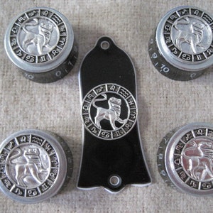 Op de afbeelding: Zes zilverkleurige knoppen met zwarte cijfers en een zwarte gitaar pickguard met een zilveren leeuw zodiac symbool. De knoppen hebben een zilveren leeuw zodiac symbool op een zwarte achtergrond.