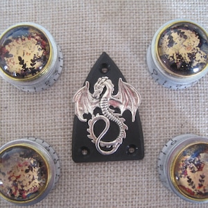 Könnte beinhalten: Vier silberne und goldene Knöpfe mit einer durchsichtigen Glasoberfläche und einem Drachenmotiv auf einem schwarzen Schlagbrett. Die Knöpfe haben die Zahlen 0-10 aufgedruckt.