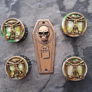 Puede incluir: Un conjunto de herrajes decorativos con temática gótica. El conjunto incluye cuatro pomos redondos con una figura de bronce dentro de un marco y una placa en forma de ataúd con una calavera y la inscripción RIP. Las piezas son de color bronce.