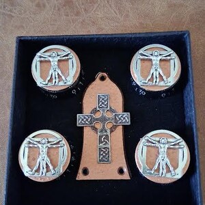 May include: A black box contains four round, silver-colored metal objects with a depiction of the Vitruvian Man, and a brown cross-shaped pendant. The items are displayed on a brown surface.