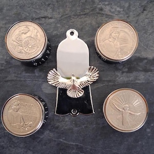 May include: A collection of silver metal knobs with black bases and coin-like tops. A silver pendant with a dove design is in the centre. The coins display various designs and text.