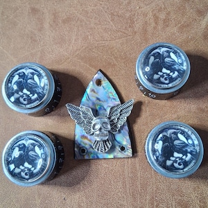 May include: A selection of guitar components, featuring four black and silver knobs with a raven motif and a decorative piece with a skull and wings. The knobs display number markings. The decorative piece has a mother-of-pearl background.