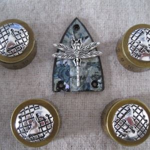 Puede incluir: Cinco cajas redondas de metal decorativas con un diseño plateado de un globo terráqueo en una cuadrícula. Las cajas están dispuestas en semicírculo alrededor de un alfiler de libélula plateado sobre un fondo azul y gris.