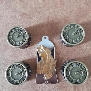 May include: Five vintage-style knobs with Roman numeral clock faces and black numbers. The knobs are silver with a gold finish. The centre knob features a decorative, gold-coloured, female figure.