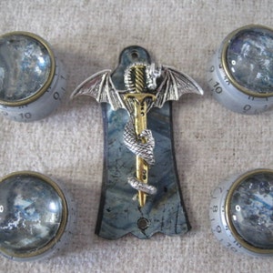 Op de afbeelding: Vijf zilveren en gouden knoppen met een blauwe en grijze gitaar pickguard met een zilveren draak en zwaard ontwerp. De knoppen hebben de nummers 0-10 erop gedrukt.