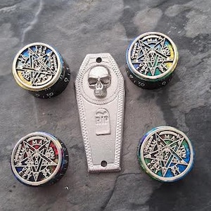 May include: A collection of gothic-themed accessories, including four round dice with pentagram designs and a silver coffin-shaped pendant with a skull and "RIP" inscription. The dice have colorful accents.