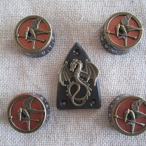 Op de afbeelding: Set van vijf gitaarknoppen met een rood en gouden ontwerp. De knoppen hebben een gestileerde vogel met een speer. De middelste knop heeft een zwarte en gouden draak.