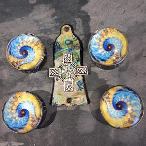 May include: A selection of handcrafted jewellery items. Features four round cabochons with a blue, yellow, and orange spiral design, and a pendant with a Celtic cross. The pendant has a pearlescent background.