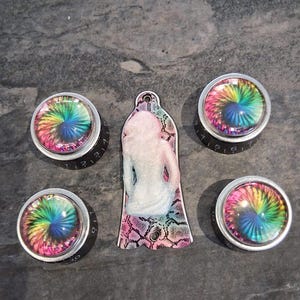 May include: A set of decorative knobs and a pendant. The knobs feature a rainbow spiral design under a clear cover, with a silver rim and black base. The pendant has a white figure on a pink and black snakeskin pattern.