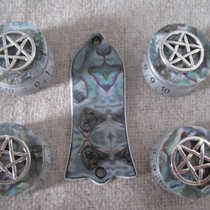 May include: A collection of guitar components: four control knobs and a switch plate. The knobs feature a silver pentagram design set against a mother-of-pearl effect. The switch plate has a matching iridescent finish.