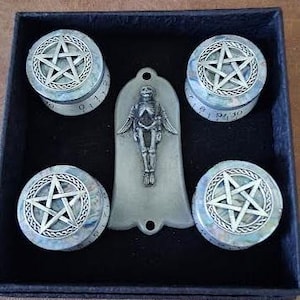 Könnte beinhalten: Eine schwarze Box enthält vier runde, hellfarbene Behälter mit einem Pentagramm-Design auf der Oberseite. In der Mitte befindet sich ein silberfarbener Skelettanhänger. Die Behälter haben ein strukturiertes, irisierendes Aussehen, und die Pentagramme sind umrandet.