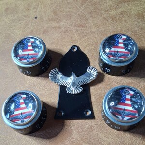 Könnte beinhalten: Eine Sammlung von Gitarrenteilen, darunter vier schwarze Knöpfe mit einem patriotischen Design, das die amerikanische Flagge und ein silbernes Adleremblem zeigt. Die Knöpfe haben weiße Zahlen von 0 bis 10. Eine schwarze Gitarrenplatte mit einem silbernen Adler ist ebenfalls vorhanden.