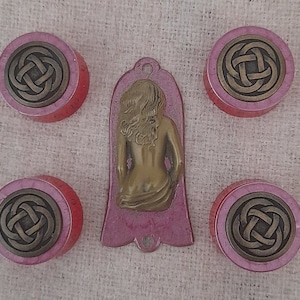 以下が含まれることがあります： ケルト結び目のデザインが施された、ピンクとゴールドの丸い装飾品が5つ。中央に女性の姿が描かれたピンクとゴールドのペンダント。