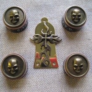 Könnte beinhalten: Satz von fünf goldfarbenen Gitarrenknöpfen mit Totenkopf-Designs. Die Knöpfe haben ein erhabenes Totenkopf-Design mit einem Kreuz und Flammen in der Mitte. Die Knöpfe sind für eine Gitarre oder ein anderes Musikinstrument.