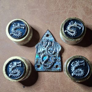 May include: A collection of fantasy-themed decorative items. The items include four round knobs with a dragon design and a triangular pendant with a dragon design. The dragon designs are silver-colored and set against a dark blue background.