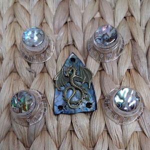 May include: Four clear plastic control knobs with numbered scales from 0 to 10 and iridescent abalone shell inlays, arranged around a triangular abalone shell guitar truss rod cover featuring a central antique brass-toned dragon emblem.