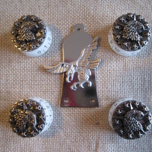 Puede incluir: Cinco pomos blancos con números negros y un diseño de águila plateada. Los pomos son para una guitarra u otro instrumento musical. Una placa de metal plateada con un diseño de águila está en el centro de la imagen.