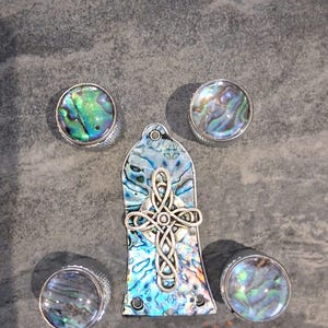 Könnte beinhalten: Gitarrenknöpfe aus Abalone-Muscheln und ein passendes Gitarrenkopfplatten-Overlay mit einem keltischen Kreuzdesign. Die Knöpfe haben einen silbernen Metallrand. Die Abalone-Muschel hat schillernde Farben von Blau, Grün und Lila.