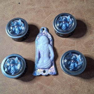 May include: A collection of decorative knobs and a pendant. The knobs have a silver rim and a blue and white image of a Norse god. The pendant is a white, seated figure with a purple and blue iridescent border.