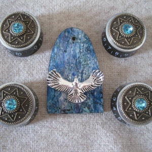 Könnte beinhalten: Vier silberne und bronzene Knöpfe mit blauen Edelsteinen und einem Sternmuster sowie eine blau-weiß bemalte Holzplakette mit einem silbernen Vogelschnitt.