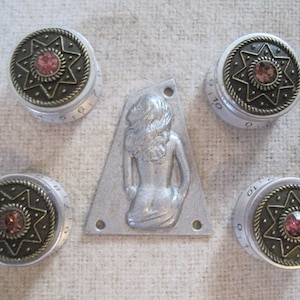 May include: Five silver and gold knobs with a star design and pink rhinestones. The knobs are surrounded by a silver trim and have numbers printed on them. A silver metal plate with a raised design of a woman's torso is in the centre.