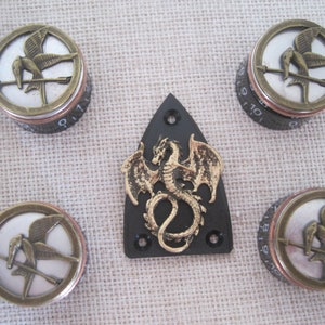 Op de afbeelding: Vijf knoppen met een gouden en zilveren ontwerp van een spotvogel met een pijl erdoorheen. Een zwarte gitaar pickguard met een gouden draak ontwerp.