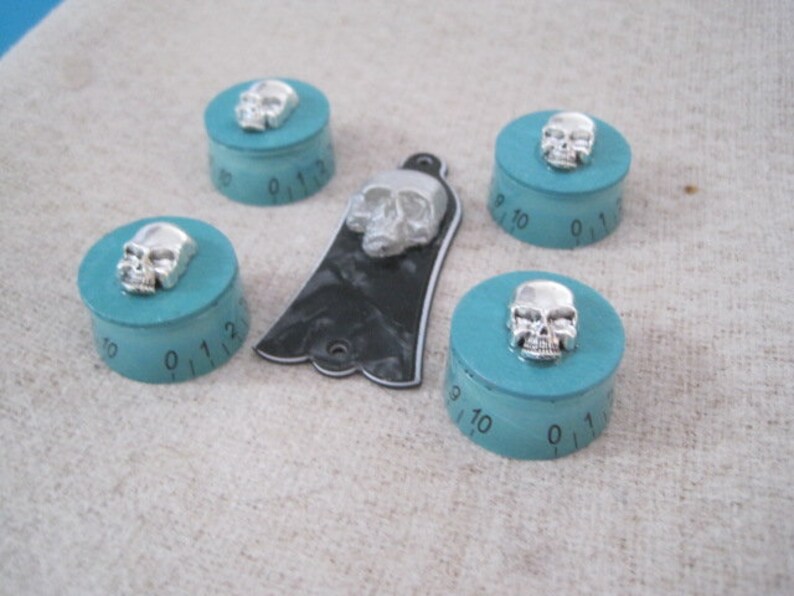 Puede incluir: Cinco pomos turquesa con detalles de calavera plateada. Cada pomo tiene un dial circular con los n&uacute;meros del 0 al 12.